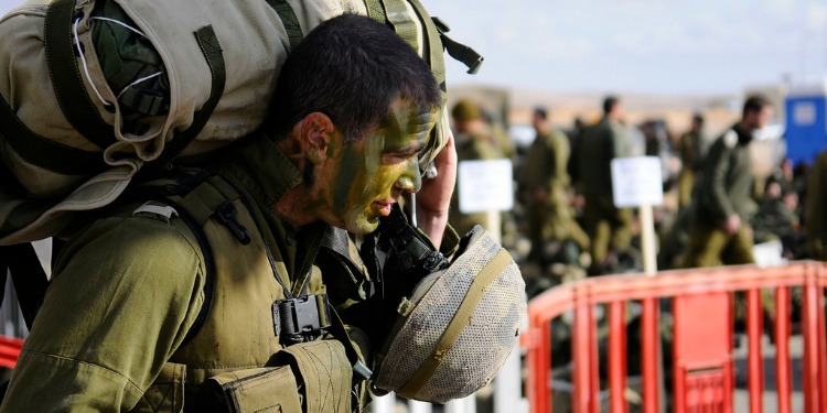 Soldier in his uniform and paint holding his helmet and backpack.