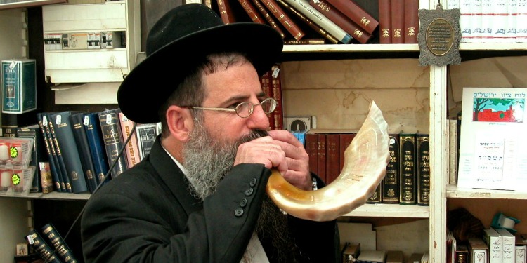 Man blowing a shofar with several bookcases behind him.