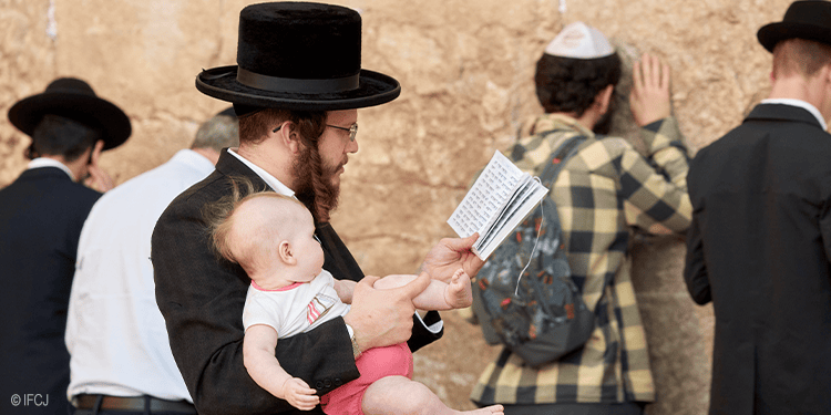 Jewish men praying at the Western Wall in Jerusalem, some reading from prayer books, others with hands on the wall, demonstrating faith and devotion.