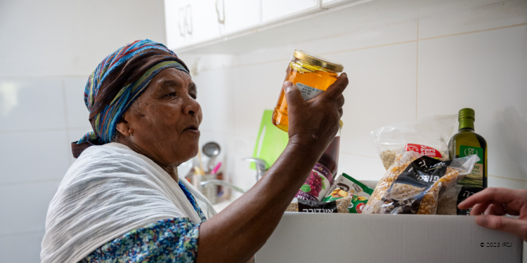 Tova Hadarai, With Dignity and Fellowship Rosh Hashanah 2023, Nof HaGalil, Israel, elderly woman with branded logo food box, holding jar of honey, floral dress, headscarf, white shawl, sad, lonely