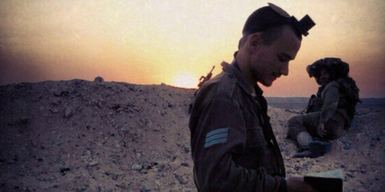 Man in uniform looking down while a man is laying down on a sandy hill behind him.