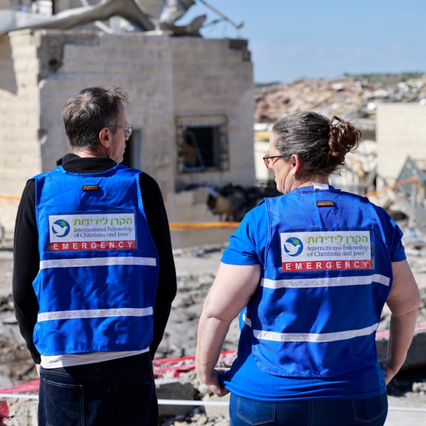 Two emergency relief workers in blue uniforms providing aid in a disaster-stricken area with damaged buildings and debris.