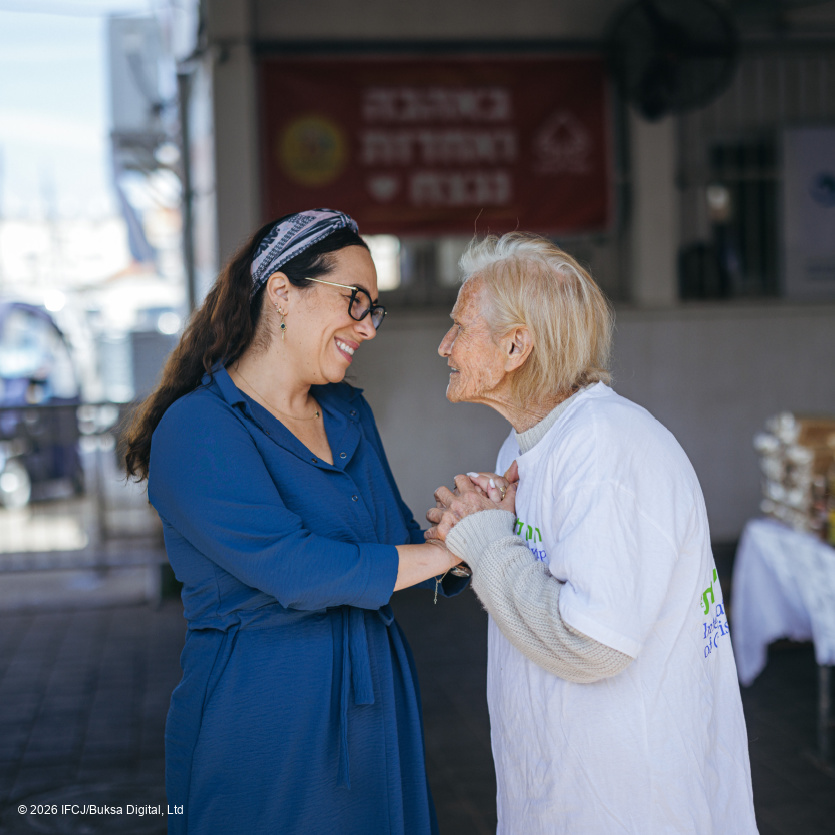 Yael Eckstein, blue dress, glasses - standing outside - talking to elderly woman, blonde hair, volunteer in branded logo t-shirt - holding hands, smiling