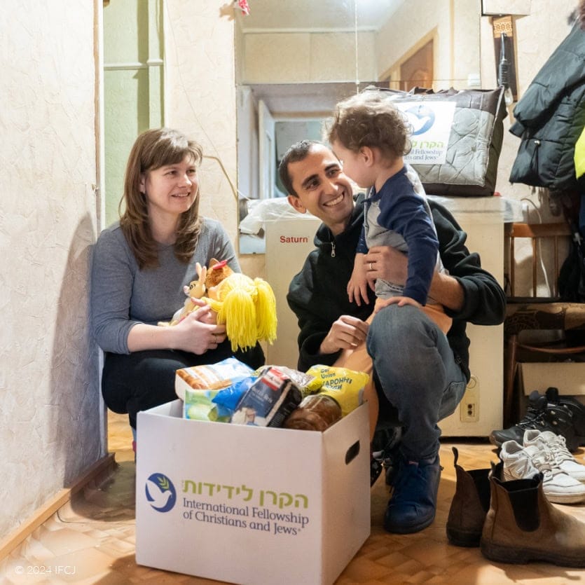 Gadi Dan (IFCJ Staff) and Yelena smiling at Carolina - black branded logo jacket - white branded logo food box - hallway - Yelena, dark gray shirt, holding stuffed doll with yellow yarn hair - Carolina, light gray shirt with navy blue sleeves, light orange pants - winter blanket in branded logo storage bag