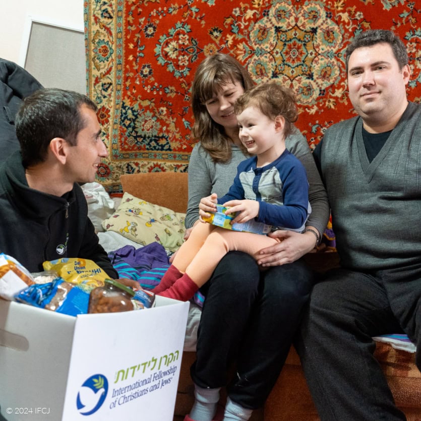 IFCJ Staff delivering white branded logo food box - black branded logo jacket - Boris, Yelena and Carolina siting on a bed - black shirt, gray sweater vest, dark gray shirt, light gray shirt with blue sleeves, orange pants, red socks - red and gold Oriental rug hanging on wall - Gadi and Carolina looking each other