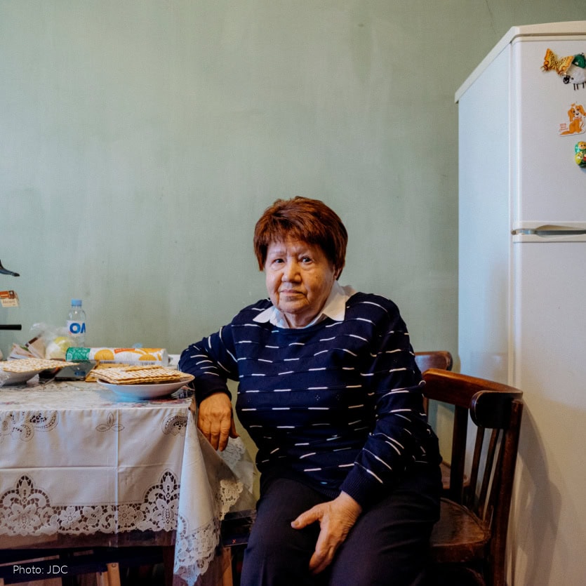 Elderly woman sitting at kitchen table - looking at camera - black and white sweater - table covered in white tablecloth with lace edge - matzah on table - white refrigerator
