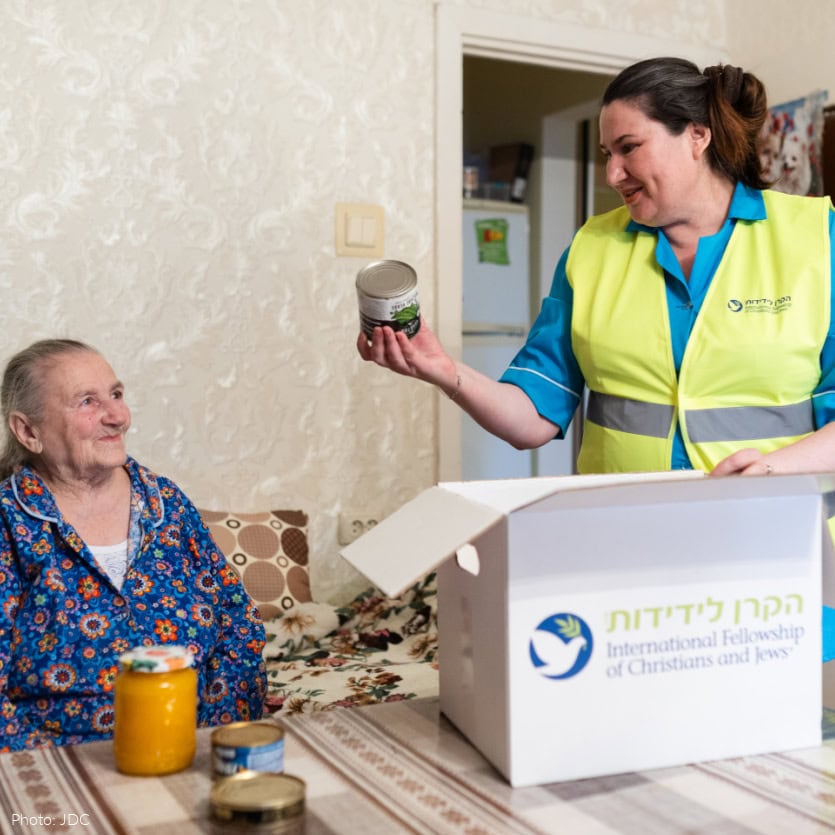 Elderly woman in blue pajamas with floral pattern, sitting on a bed - woman in turquoise blue shirt, yellow safety vest - opening branded logo food box on table with brown and beige tablecloth - holding up canned food