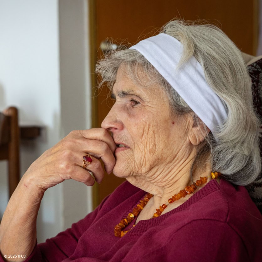 Elderly woman with gray hair, wearing a white headscarf and red sweater, sitting indoors with a contemplative expression, representing compassion and support for seniors.