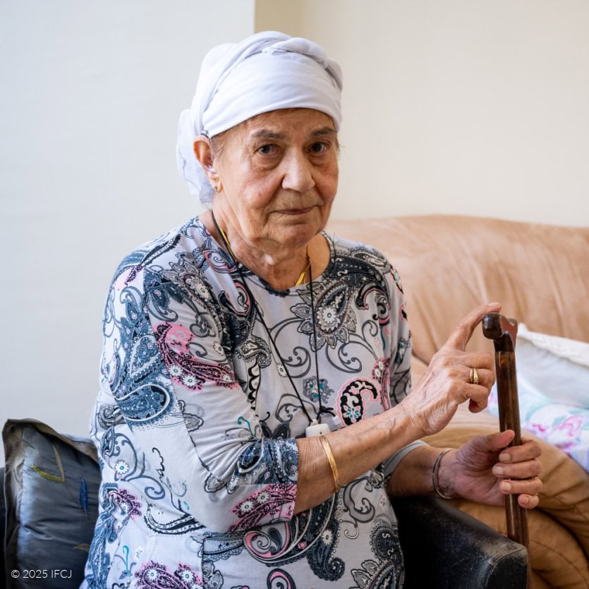 Marcel Bar David visit to elderly woman's home. elderly woman, blue and green paisley dress, headscarf, sitting in black armchair, holding cane. Brown couch, orange blanket, white pillows