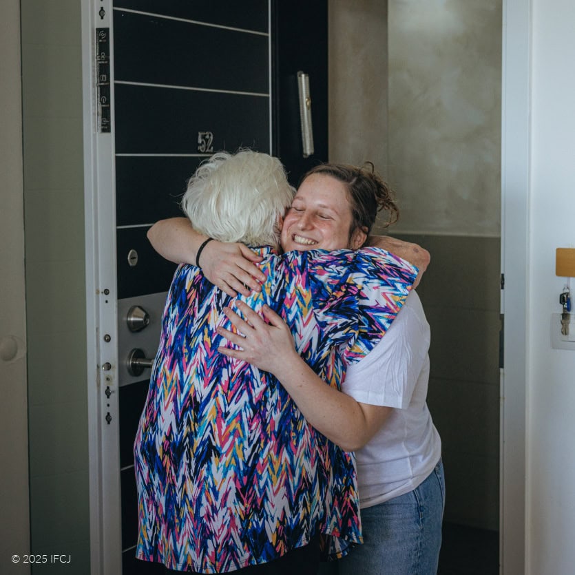 Elderly woman opening door, seen from behind - greeting IFCJ staff woman in open door - blue and pink patterned blouse, white T-shirt with IFCJ logo - smiling - hug
