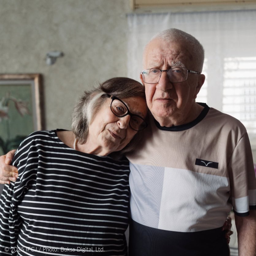 Moshe Adler - elderly man, glasses, tan, gray shirt, standing - with his wife, Malka Adler - elderly woman in black and white striped shirt - arms around shoulder - gray couch - purple flowers - her head on his shoulder
