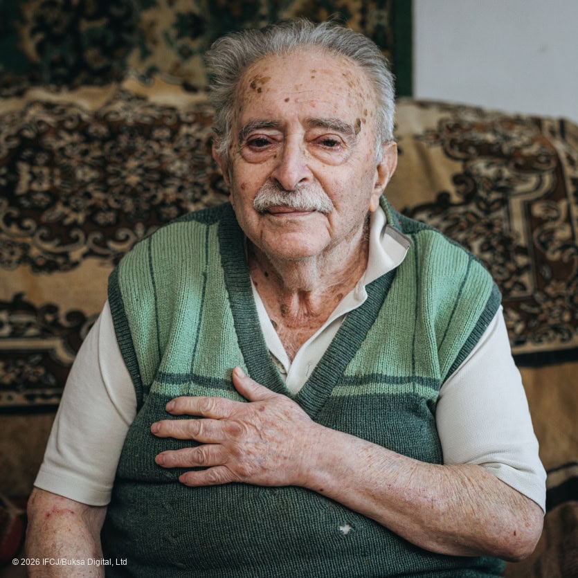 An elderly man with gray hair and a mustache sitting on a sofa, wearing a green sweater, in a cozy home setting, representing care and compassion.