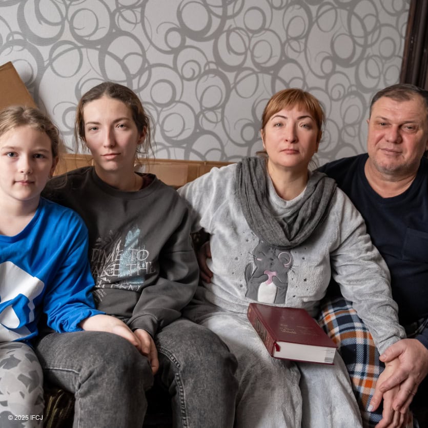 Family sitting on couch - 2 adult women, adult man in plaid pants, girl in blue and white shirt, gray swirls wallpaper, carboard boxes flattened - woman holding Torah book on her lap