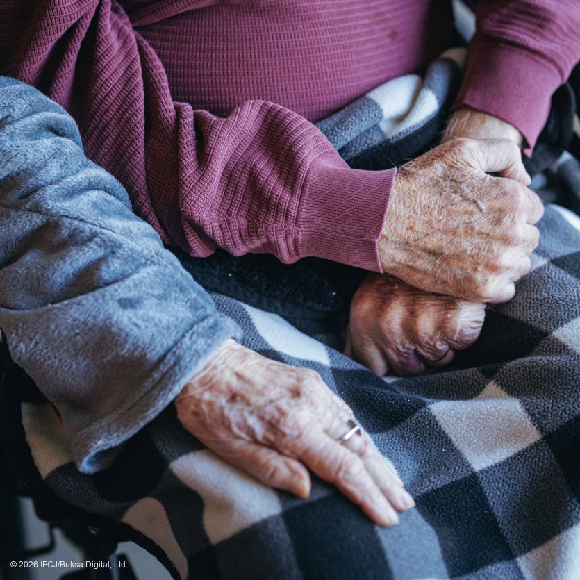 Dalia, woman in blue jacket - Pinhas, elderly man, purple long-sleeved t-shirt, black and white buffalo check blanket on his lap - her hand on his knee - no faces, focus on hands