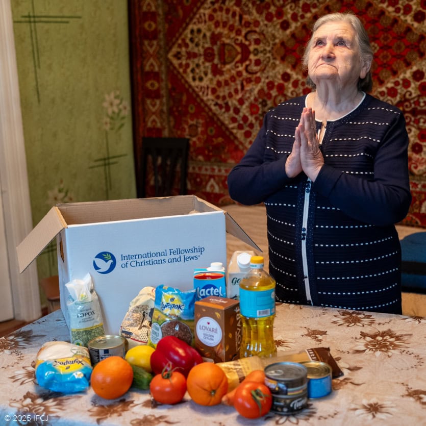 Elderly woman, black and white dress, looking up, holding hands in prayer - IFCJ logo food box on table with tan tablecloth - Oriental rug on wall - green wall - oranges, red pepper, milk, produce