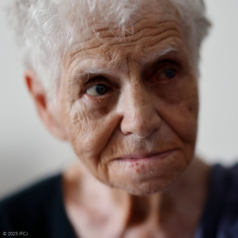 Close-up of elderly woman with short white hair, facing slightly to the side, with a softly furrowed brow, downturned mouth, and a steady gaze away from the camera, set against a softly lit, neutral indoor background.