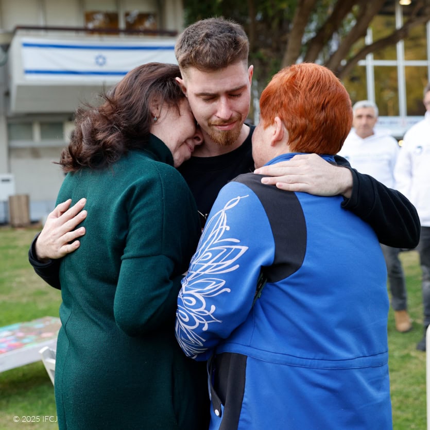 Vladimir Matveychuk and Svetlana Matveychuk - Tel Aviv, Israel - February 26, 2025 Injured Lone Soldier Visited by Ukrainian Mother and Aunt young adult man, black sweatsuit, outside, green grass, hugging adult woman with short red hair, light blue jacket, and adult woman with dark hair, dark green turtleneck dress, emotional - IFCJ staff standing in background, watching