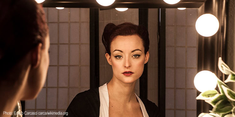 Portrait of a woman with red hair and fair skin, wearing a black and white outfit, in a traditional Japanese-style room with shoji screens and warm lighting.