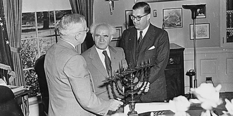 Black and white photo of three men talking over a menorah.