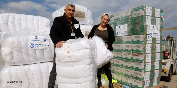 Yael Eckstein and an IFCJ coworker holding blankets with boxes of blankets behind them.