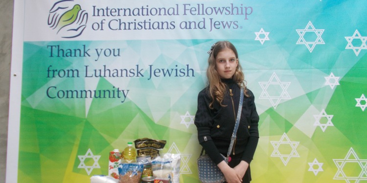 Alika, young IFCJ recipient standing in front of an IFCJ branded sign with her food box contents.