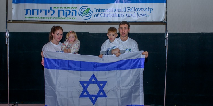 IFCJ recipient family holding the Israeli flag in front of an IFCJ branded banner.