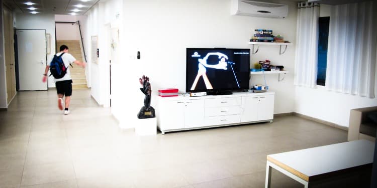A teenager walking through a white room with a table, chair, TV, TV stand, and games.
