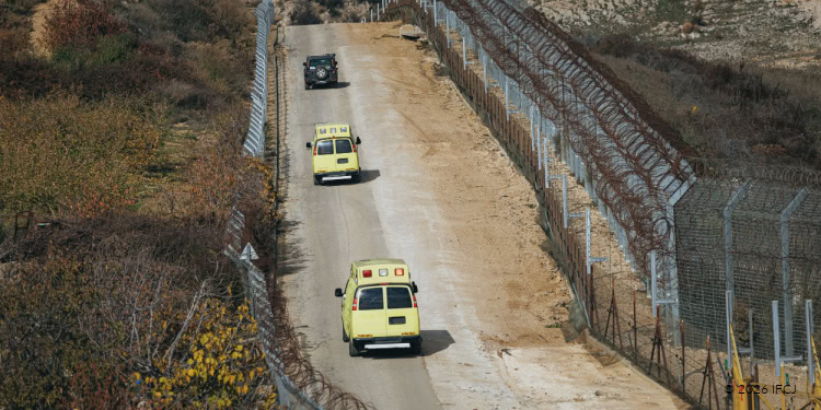 Two ambulances driving along a dirt road surrounded by barbed wire fences and dry vegetation, highlighting humanitarian efforts in conflict zones.