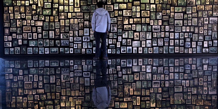 Man stands in front of wall full of photos of victims of the Holocaust.