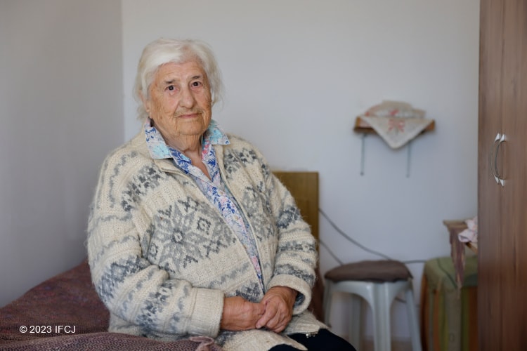 Elderly Jewish woman in white sweater sitting on her bed while looking into the camera.