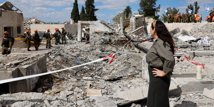 Yael Visits Beit Shemesh - Iran attacks / Operation Roaring Lion (Epic Fury) - March 8, 2026 Yael Eckstein, gray blouse, black skirt - site of a missile hit March 1, 2026 - surrounded by debris, destroyed buildings - people in orange safety helmets in background