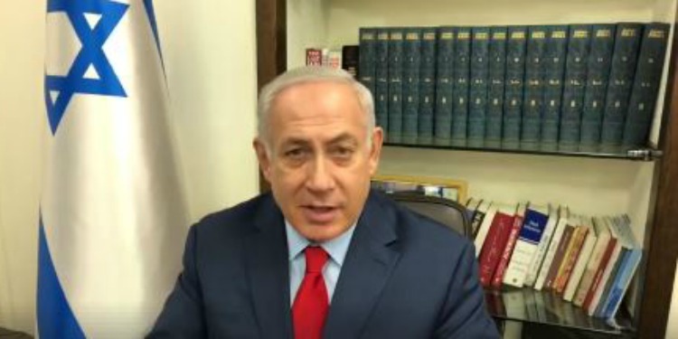 Bibi giving an interview in a library with books and an Israeli flag beside him.