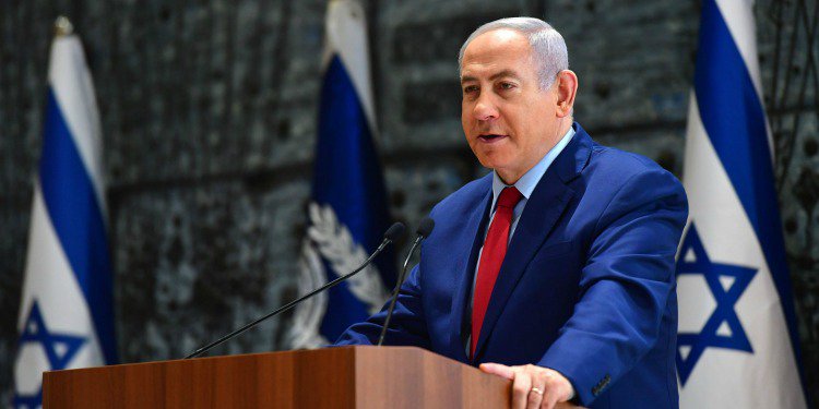 Bibi in a suit at a podium speaking while Israeli flags are behind him.