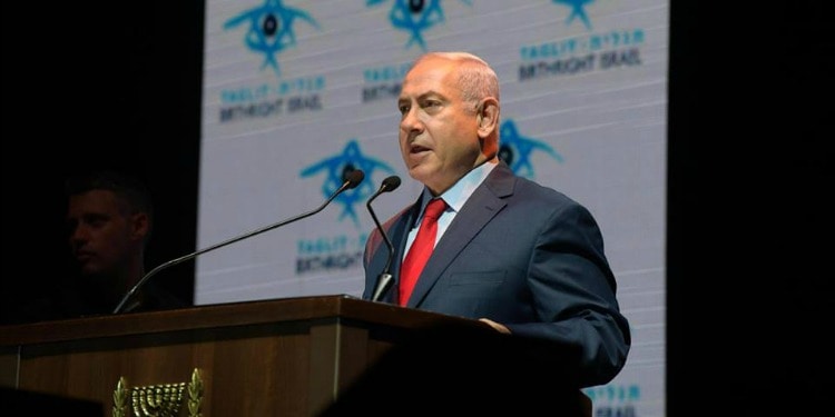Bibi giving a speech at a podium with a blue and white patterned banner behind him.