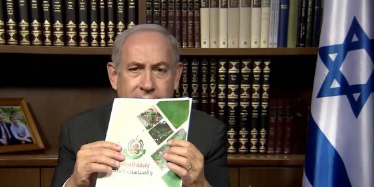 Bibi holding a pamphlet while sitting in a library with the Israeli flag beside him.