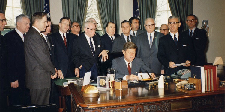 JFK and his cabinet standing behind him in the Oval Office.