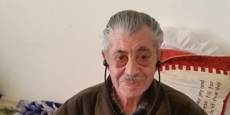 Elderly Jewish man with headphones in sitting on a bed.