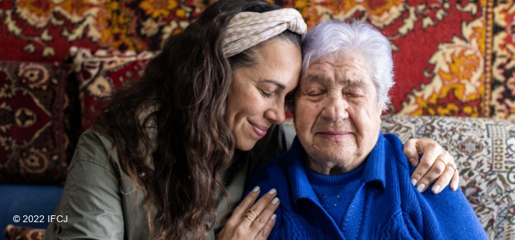 Yael Eckstein hugging Bronya Liftziger, an elderly Jewish woman while they sit on a couch together.