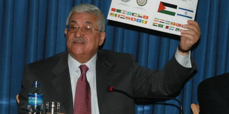 Elderly man at a podium on a stage holding a piece of paper with flags on it.