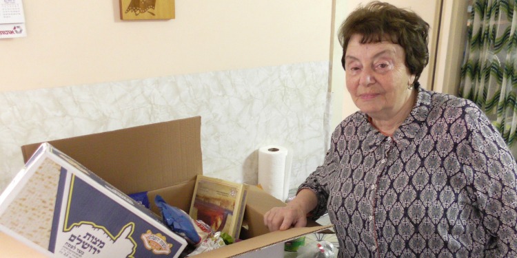 Elderly Jewish woman standing next to an IFCJ branded food box delivered to her home.