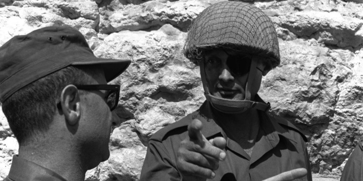 A soldier with a helmet and an eye patch on pointing forward as he's talking to another soldier.