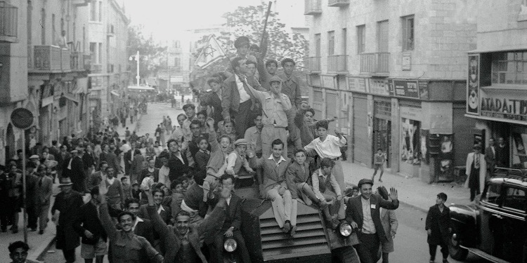 Several men and boys on top of a car waving the Israeli flag and yelling.