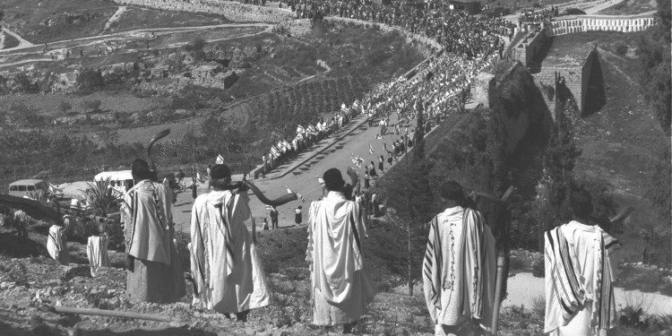 Black and white image of men on a hill with shofars while a parade is going on below them.