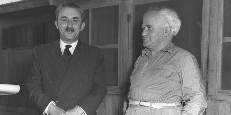 Two men, one of them Moshe Sharett standing outside of a wooden building.