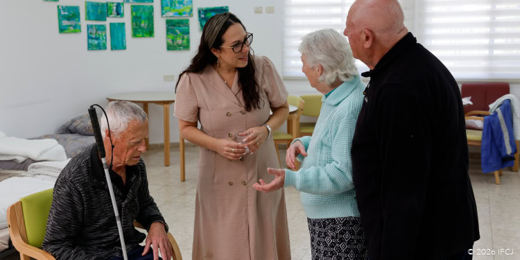 Visit to Elderly Day Center - Filming in Dimona, Israel - March 30, 2026 Yael Eckstein, beige coat dress, glasses - standing, talking to Joseph and Anna Zarkhovsky - elderly man, black shirt, elderly woman, light green sweater - elderly man, seated, white cane, blind - listening