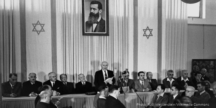 Group of people celebrating Yom HaAtzmaut in Israel, with a speaker at the podium and Israeli flags, commemorating Israel's Independence Day.