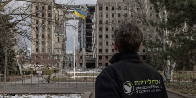 Gidi Shmerling (IFCJ Staff) standing in front of destroyed building in Mykolaiv, from behind, Ukraine flag on flagpole.