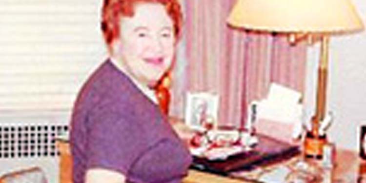 A middle-aged woman in a purple top with red hair smiling while sitting at a desk in her home.