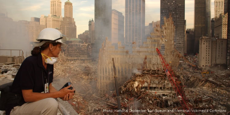 Rescue worker in protective gear at the 9/11 site with damaged buildings and smoke in the background, highlighting the aftermath of the September 11 attacks.