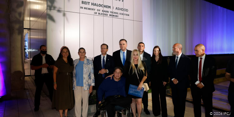 Group of people at the Beit HaLochem Soldiers Home opening event, including officials and community members, in front of a white wall with the IFCJ logo.